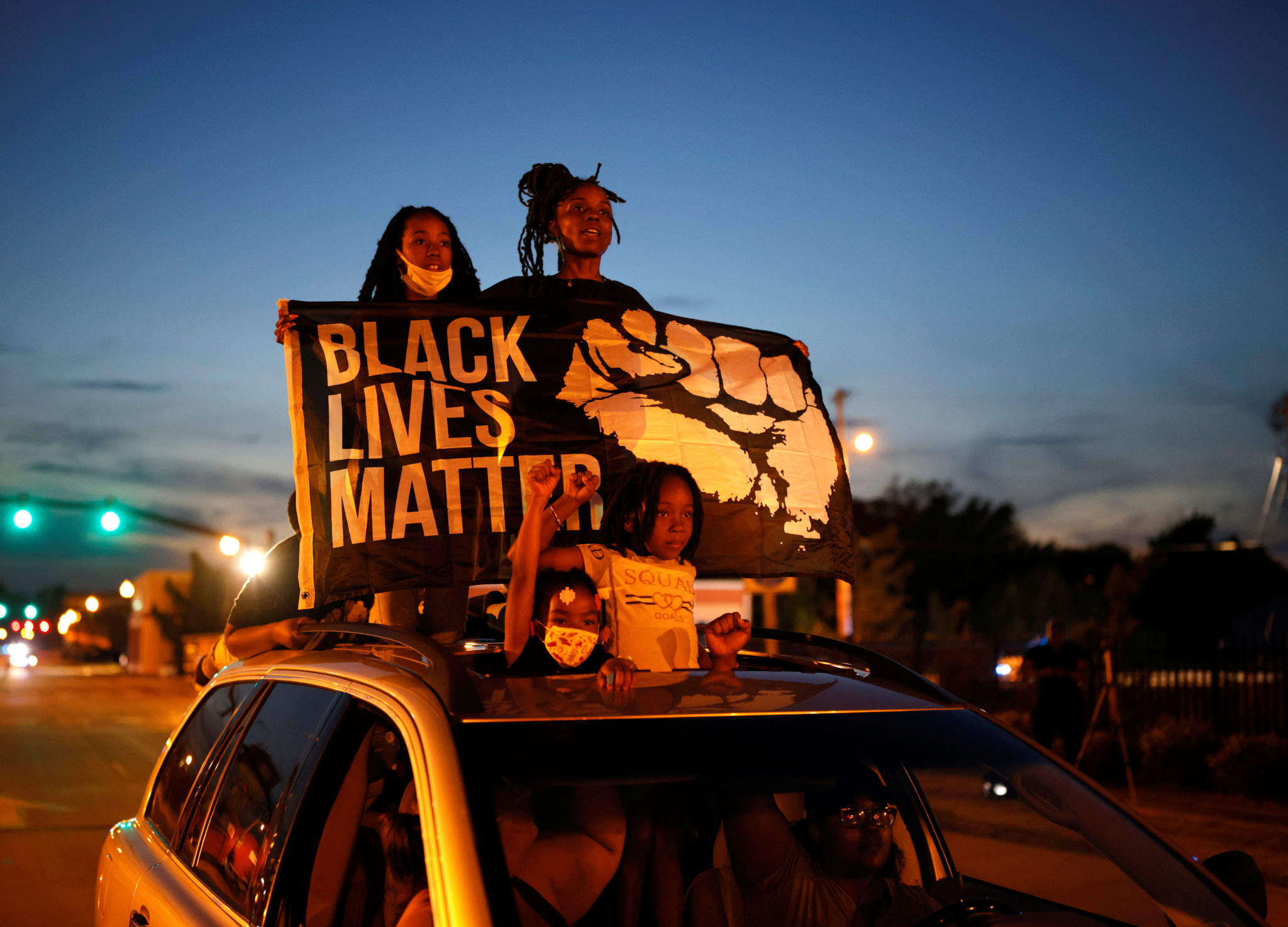 A group of Black protesters stand through the sunroof of a car with a large protest banner which reads 'Black Lives Matter' with a Black power fist.