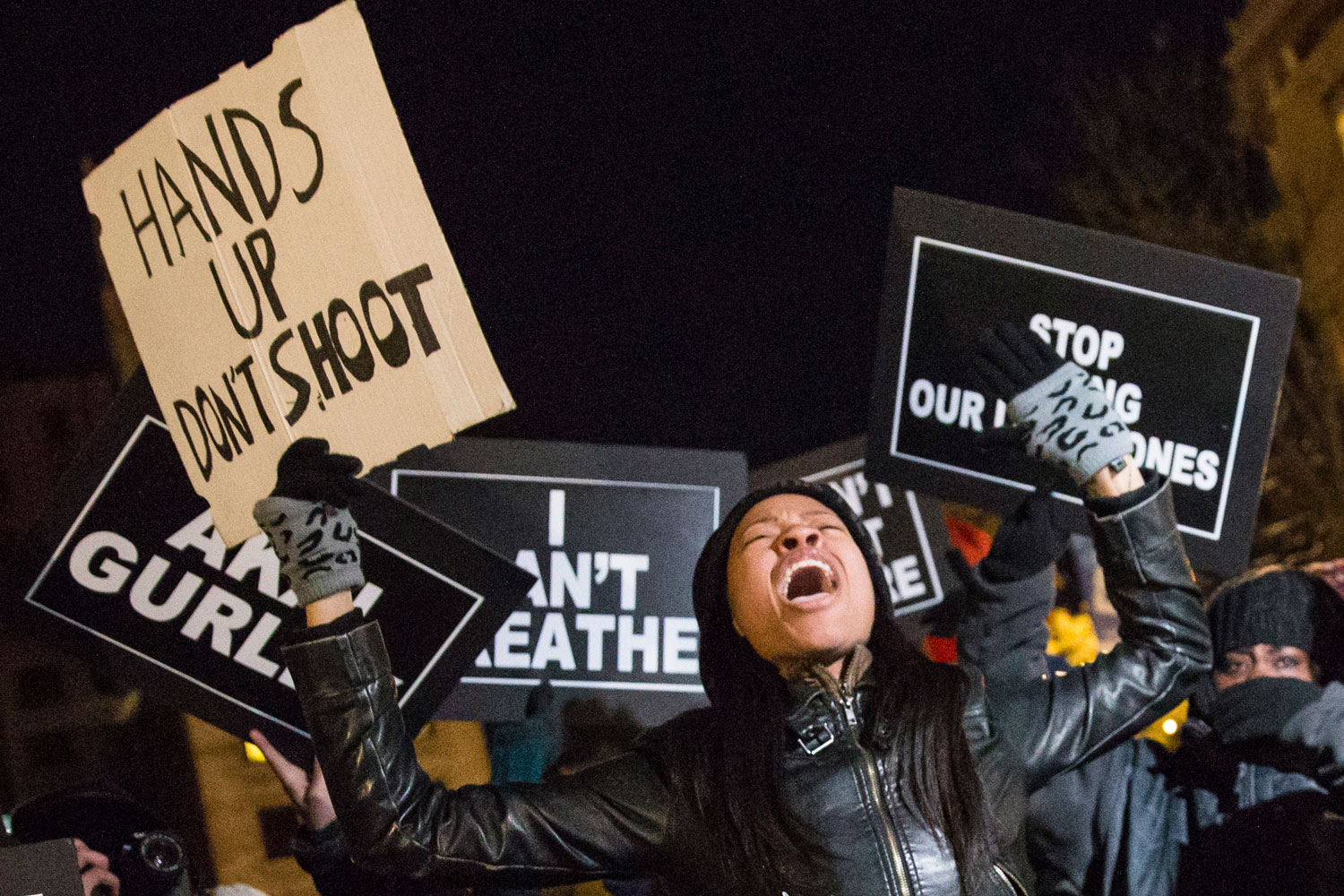 A Black female protester chants and raises her hands in the air while holding a sign that reads, 'HANDS UP DON'T SHOOT.'